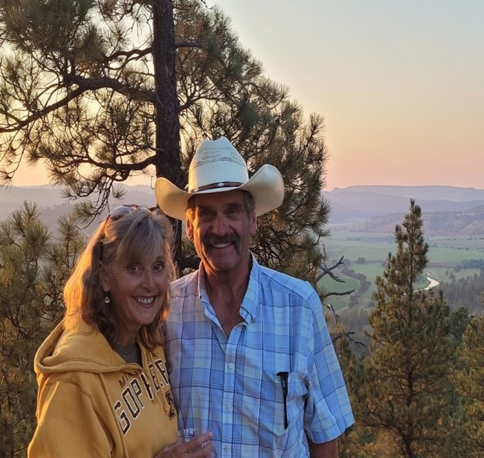 A woman and man stand in front of a tree with a scenic backdrop.