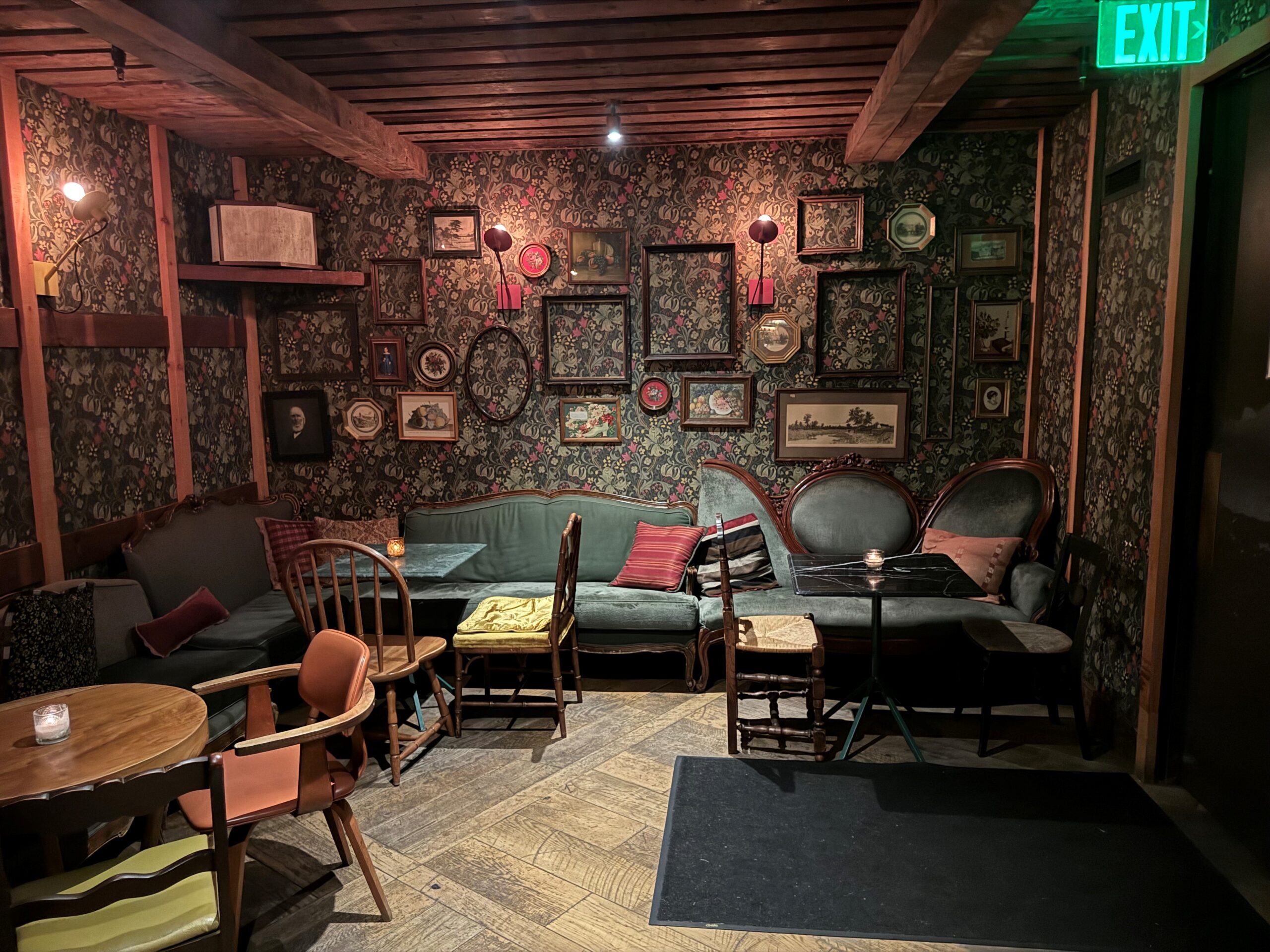 A trendy speakeasy area showing green couches and wood tables in front of a wall, covered in wallpaper with empty frames.