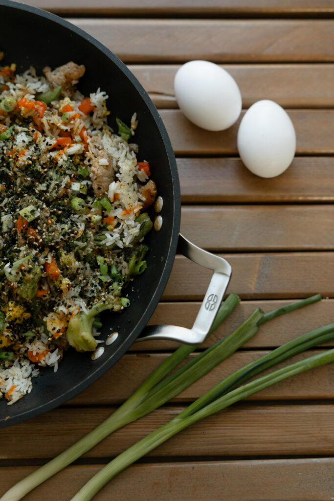 Two eggs and two green onions alongside a frying pan full of pork fried rice.